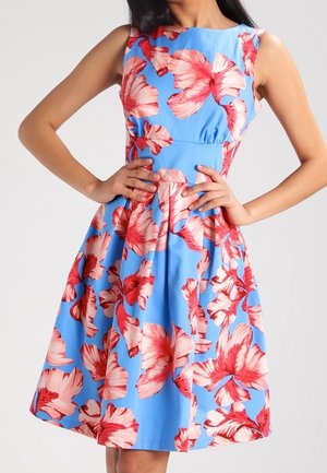 Woman wearing a sleeveless blue dress with large red and pink floral patterns, hands resting on hips, standing against a plain background.