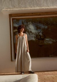 Gestreiftes Maxikleid in Creme und Beige mit Rüschenträgern; kombiniert mit blauen und braunen offenen Schuhen. Sonniger Hintergrund.