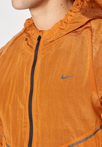 Oranje lichtgewicht jack met capuchon, voorzien van een zwarte rits, grijs accentlogo en een gerimpelde textuur. Reflecterende strip langs de onderrand.