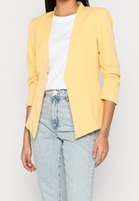 Femme portant un blazer jaune pâle à manches froncées sur un t-shirt blanc et un jean taille haute bleu clair, debout les bras détendus.