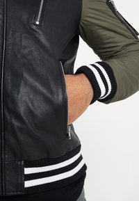 Veste aviateur en cuir noir avec manches vert olive. Dotée de poignets côtelés noir et blanc, d'une fermeture éclair à l'avant et d'une poche latérale zippée.