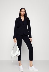 Costume noir avec une veste ajustée et un pantalon slim, associé à des baskets blanches. Un sac à main blanc est tenu d'une main. Tissu lisse, design structuré.