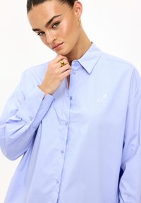 Camisa de botones azul claro con cuello, con mangas largas y un logotipo bordado de 'L' en el pecho. Tejido suave y corte relajado.