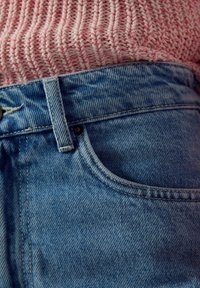 Primo piano della vita di jeans blu con passante per cintura, parzialmente coperta da un maglione rosa lavorato a maglia.