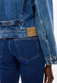 Giacca in denim con una colorazione blu sbiadita e accessori in ottone, con un'etichetta in pelle marchiata. Jeans scuri attillati con un classico design a cinque tasche.