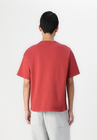 Tricou roșu din bumbac, cu mâneci scurte și guler rotund, având o croială lejeră și o textură plaină a materialului. Purtați-l cu pantaloni de jogging gri.