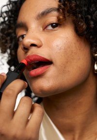 Rossetto rosso applicato, caratterizzato da una texture opaca e liscia. Il prodotto presenta un elegante packaging nero con una punta colorata. La pelle mostra toni naturali.