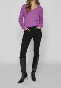 Pull violet à col en V, jean noir ajusté, et bottes en cuir noires montantes jusqu'aux genoux avec un bout pointu et un talon stiletto. Textures lisses sur l'ensemble.