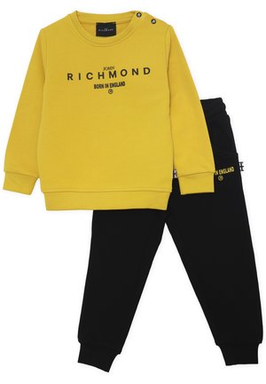 Felpa gialla con testo nero, collo rotondo e dettagli con bottoni, abbinata a pantaloni jogger neri con dettagli su tasche e logo.