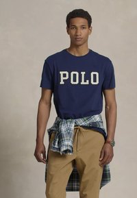 Mlad moški v mornarsko modri "POLO" majici, bež hlačah, karirasti srajci, zavezani okoli pasu, in s wristwatchom, stoji pred enobarvno ozadjem.