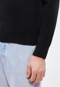 Μαύρο πλεκτό πουλόβερ με ριμπ μανσέτες, συνδυασμένο με ανοιχτό μπλε τζιν παντελόνι. Διαθέτει μικρή λεπτομέρεια με λογότυπ�ο κοντά στο τελείωμα. Η υφή φαίνεται απαλή.