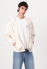 Beige zip-up hoodie med snoretræk, frontlommer, ribbede manchetter, båret over en hvid T-shirt, parret med lyseblå jeans.