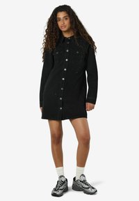 Žena s kudrnatými vlasy oblečená v černých džínových šatech na knoflíky se zdobením, bílými ponožkami a stříbrno-černými teniskami na bílém pozadí.