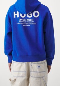 Felpa blu in pile con un prominente logo e testo bianco "HUGO" sul retro. Presenta una tasca a marsupio e un cappuccio regolabile con coulisse.
