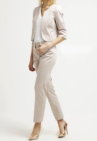 Femme portant un blazer court beige clair, un pantalon tailleur assorti, un chemisier blanc et des chaussures à talons hauts métalliques dorées, debout de profil.