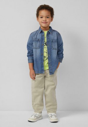 Junger Junge steht mit den Händen in den Taschen, trägt ein blaues Jeanshemd, ein hellgelbes T-Shirt mit Grafik, beige Hose und weiße Sneakers vor grauem Hintergrund.