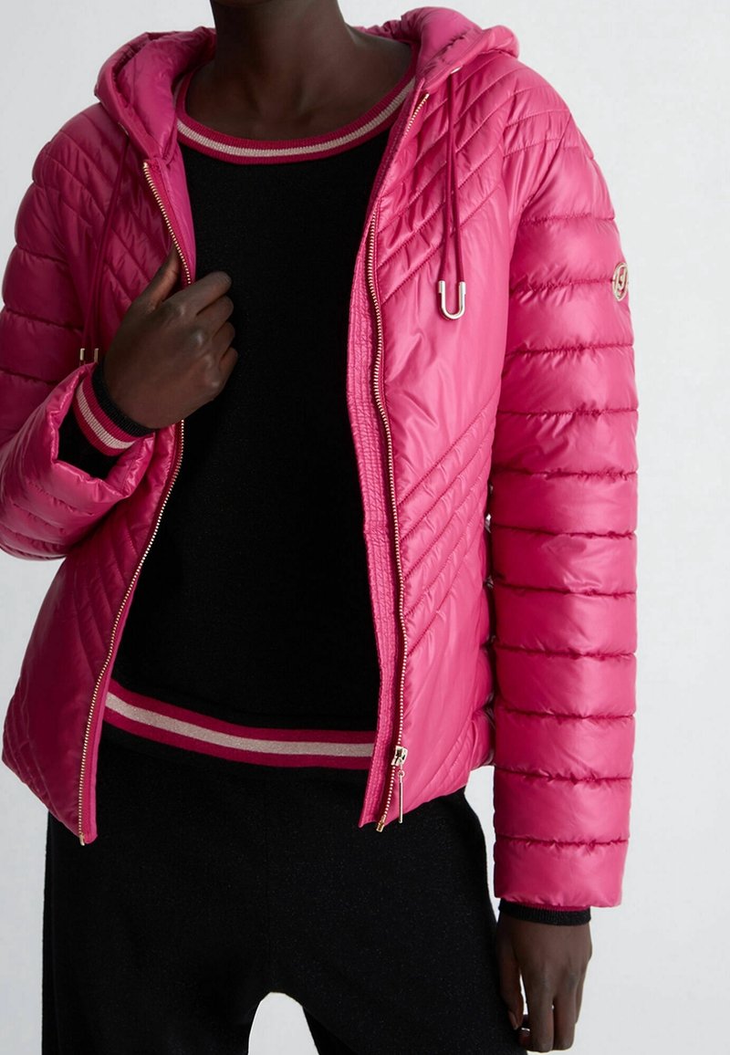 Personne portant une veste matelassée rose vif avec capuche et fermeture éclair partiellement ouverte, par-dessus un pull noir avec des bordures rayées rose et blanc.