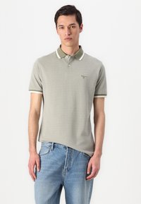 Polo cinza claro com gola e punhos em listras verdes escuras e brancas, apresentando um pequeno logotipo no peito, usado com jeans azuis.