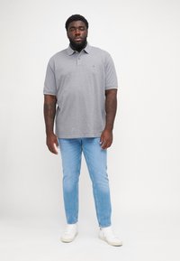 Polo grigio a maniche corte, con colletto, abbinato a jeans azzurri chiari e sneakers bianche. Presenta un piccolo logo sul lato sinistro del petto.