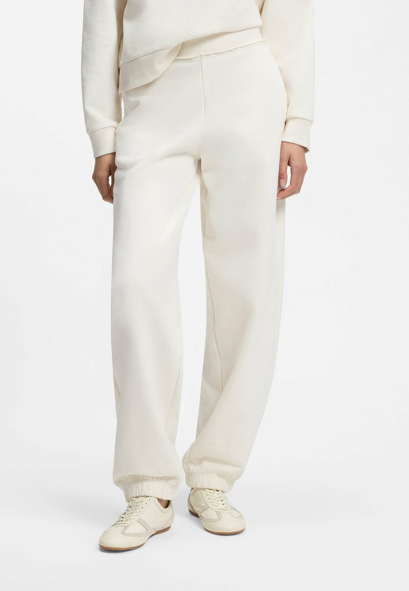 Personne portant un pantalon de survêtement crème avec des poignets élastiques, des baskets crème assorties et un sweat-shirt crème sur un fond uni.