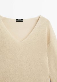 Beige gebreide trui met een V-hals, met een zachte textuur en een losvallend ontwerp. Label zichtbaar bij de halslijn.