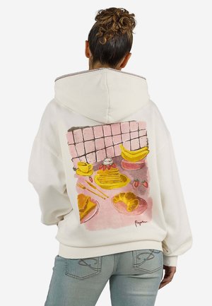 Person med krøllet hår i en knold har en hvid hættetrøje på med en farverig morgenmadillustration af pandekager, croissant, bananer og kaffe på ryggen.