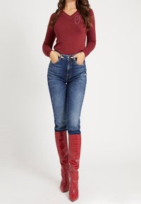 Haut bordeaux à manches longues avec col en V et coutures décoratives, assorti à un jean bleu taille haute et des bottes rouges brillantes montant jusqu'aux genoux.