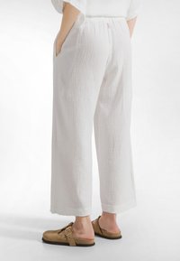 Henkilö, joka pukeutunut löysiin valkoisiin, teksturoiduista kankaista valmistettuihin housuihin, joissa on sivutaskut, sekä beigenruskeisiin mokkanahkaisiin liukusukkakenkiin, joissa on solkikoristelu, kuvattuna takaa.