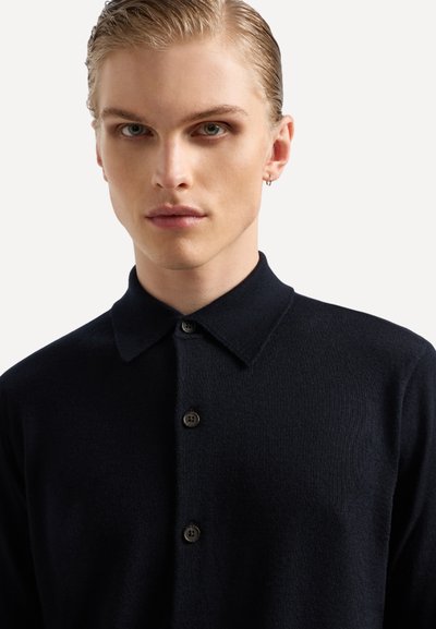 Polo in maglia blu navy con colletto con bottoni, presenta cinque bottoni marroni e una texture morbida. Vista ravvicinata che mette in risalto il design e il colore.