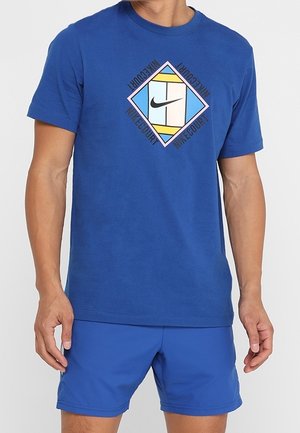 Homme portant un t-shirt Nike bleu avec un logo carré coloré Nike Court et un short bleu assorti, debout les bras détendus.