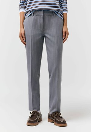 Osoba ubrana w szare spodnie w prążki, brązowe sznurowane buty oraz niebieską koszulę w paski z długim rękawem, stojąca na białej podłodze.