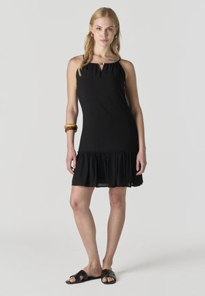 Mujer de pie vistiendo un vestido negro sin mangas con dobladillo con volantes, pulseras doradas en la muñeca izquierda y sandalias negras tipo slip-on sobre un fondo gris.