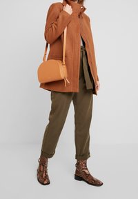 Bruine wollen jas, groene high-waisted broek en bruine leren enkellaarsjes met slangenpatroon. Draagt een kleine oranje leren crossbody tas.