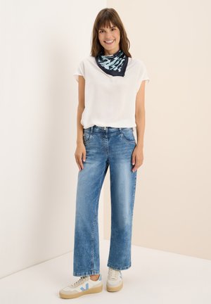 Weiße Kurzarmbluse, hellblaue weite Jeans und cremefarbene Sneaker mit blauen Akzenten; dunkel gemusterter Schal um den Hals gebunden.
