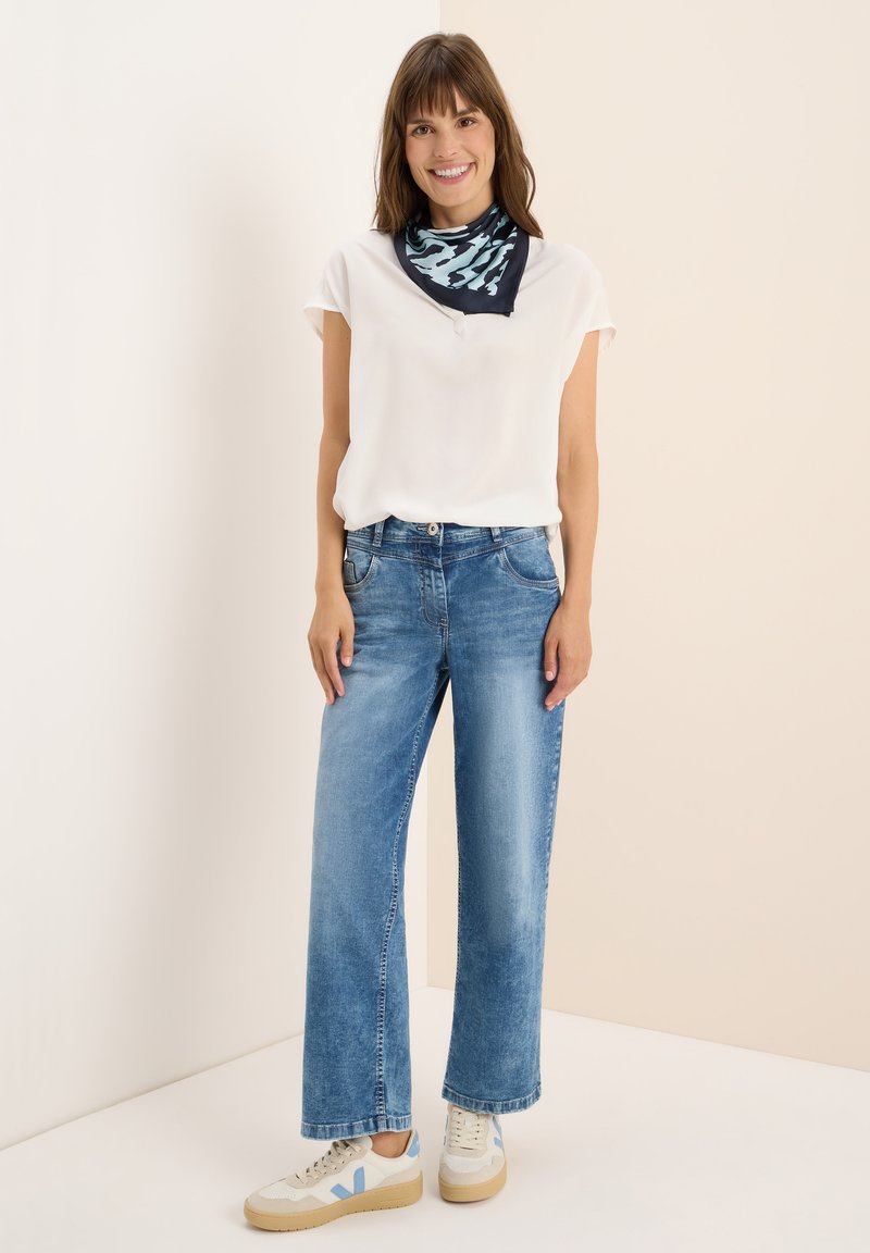 Blusa bianca a maniche corte, jeans a gamba larga blu chiaro e sneakers color crema con accenti blu; sciarpa scura a fantasia annodata attorno al collo.