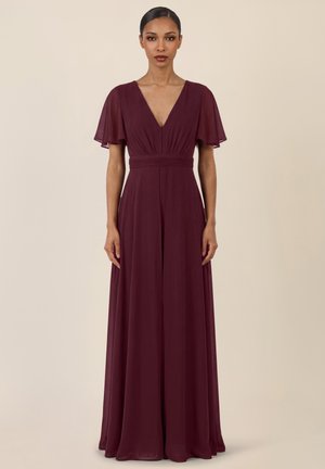 Kvinde stående iført en gulvlængde burgundy kjole med flagrende ærmer og dyb V-udskæring mod en ensartet beige baggrund.