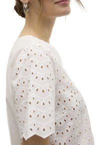 Haut en coton blanc avec broderie œillet dans un motif géométrique, doté de manches festonnées et d'une coupe ample.