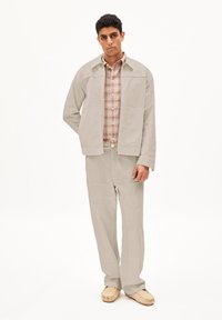 Giacca beige chiaro su misura con taglio dritto, abbinata a una camicia a motivi scozzesi leggera e pantaloni abbinati. Texture morbida e design minimale.