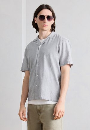 Νέος άνδρας με γκρι πουκάμισο με κοντά μανίκια πάνω από λευκή μπλούζα, μπεζ παντελόνι και σκουρόχρωμα γυαλιά ηλίου, στέκεται μπροστά σε έναν φωτεινό τοίχο.