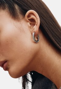 Mustast materjalist rõngaskõrvarõngas, mille välisküljel on reas selged rhinestones-kivid, kantud mudeli kõrvas ja näidates selle kaarduvat disaini.