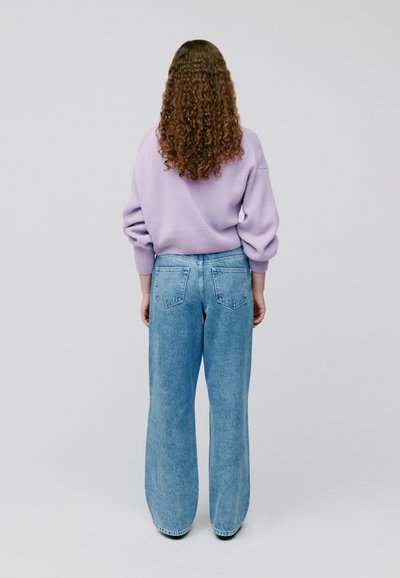 Lila übergroßer Pullover mit gerippten Bündchen, kombiniert mit hoch taillierten, hellblauen weit geschnittenen Jeans. Lockige Haare fallen über die Schultern.