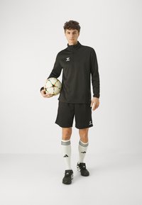 Schwarzes langärmeliges Sportoberteil, passende Shorts, weiße kniehohe Socken mit schwarzen Streifen, hält einen Fußball mit bunten Akzenten.