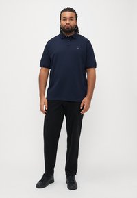 Marineblå polo med klassisk krave, korte ærmer og et lille logo på brystet, kombineret med sorte bukser og sorte sneakers.