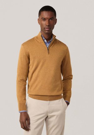 Suéter de color amarillo dorado con cuello de media cremallera, puños acanalados y dobladillo, superpuesto a una camisa a rayas azul y blanca, combinado con pantalones beiges.