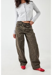 Kobieta w szarej, skróconej, zapinanej na guziki bluzie, brązowych dżinsach w leoparda o szerokich nogawkach i czerwonych balerinach, stojąca na tle białym.