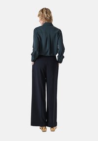 Dunkelgrüne Satinbluse mit Kragen, kombiniert mit weiten, navyblauen Hosen und metallisch goldenen Schuhen. Sichtbare Gesäßtaschen an den Hosen.