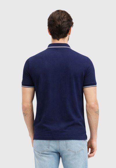 Άνδρας με κοντά σκούρα μαλλιά φοράει μπλε πόλο μπλουζάκι με λευκές λεπτομέρειες και ανοιχτόχρωμο τζιν, στέκεται με πλάτη σε απλό φόντο.