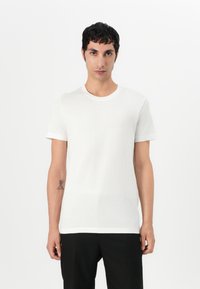 Camiseta branca de manga curta, feita de tecido leve, com um decote redondo e uma textura discreta. Usada com calças pretas, sem padrões ou enfeites.