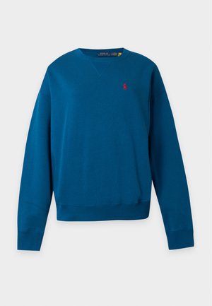 Kvinde iført mørke solbriller, blå sweatshirt, sorte bukser og hvide sneakers, står med hænderne i lommerne på en ensfarvet hvid baggrund.