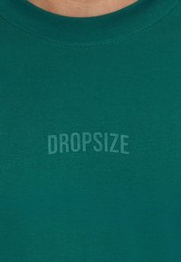 Zelena bombažna majica s kratkimi rokavi in okroglim izrezom, z rahlo embosiranim logotipom "DROPSIZE" čez prsi. Gladka tekstura in sproščen kroj.